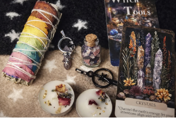 Witch tools including a rainbow-colored smudge stick, crystals, and tarot cards on a dark fabric background.